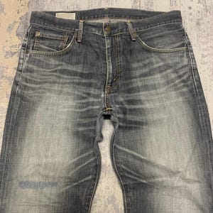 Pode incluir: Jeans de ganga azul desbotada com um design cl&aacute;ssico de cinco bolsos. Os jeans t&ecirc;m fecho de bot&atilde;o e z&iacute;per, presilhas para cinto e uma lavagem clara com desbotamento sutil e marcas de desgaste.