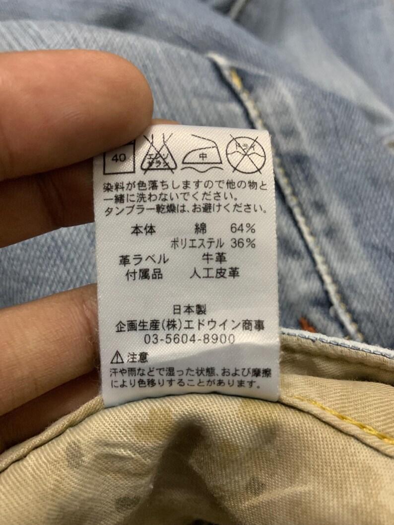 Puede incluir: Primer plano de una etiqueta con texto japon&eacute;s y s&iacute;mbolos de cuidado. La etiqueta indica que la composici&oacute;n del tejido es 64% algod&oacute;n y 36% poli&eacute;ster. Tambi&eacute;n menciona que el producto est&aacute; fabricado en Jap&oacute;n y proporciona un n&uacute;mero de tel&eacute;fono.