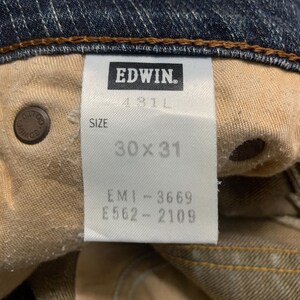Op de afbeelding: Close-up van de binnenkant van een blauwe spijkerbroek. Het label luidt "EDWIN" en geeft een maat van 76x79 cm aan. De jeans heeft een bruin gestikte zoom en een knoop met de merknaam.