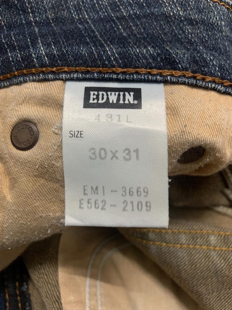 Op de afbeelding: Close-up van de binnenkant van een blauwe spijkerbroek. Het label luidt "EDWIN" en geeft een maat van 76x79 cm aan. De jeans heeft een bruin gestikte zoom en een knoop met de merknaam.