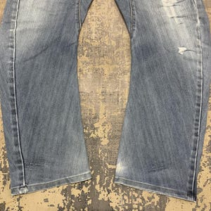 Puede incluir: Vaqueros de mezclilla azul claro con detalles desgastados. Los vaqueros tienen un dise&ntilde;o cl&aacute;sico de cinco bolsillos, una bragueta con cremallera y un corte recto. La mezclilla tiene un lavado descolorido y sutiles bigotes.