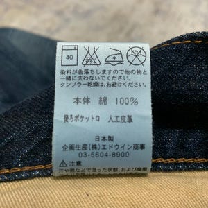 May include: Close-up of a dark blue denim fabric with a white tag attached. The tag features Japanese text and care symbols, including a washing instruction of 40 degrees. The fabric is 100% cotton.
