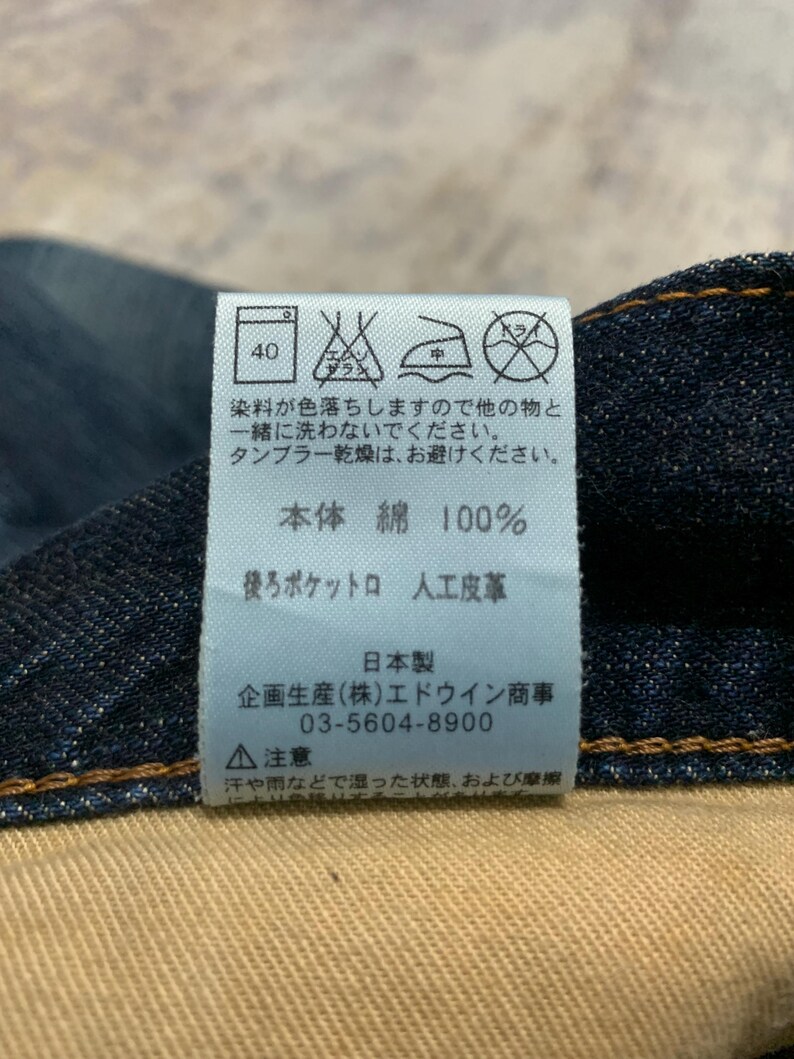 May include: Close-up of a dark blue denim fabric with a white tag attached. The tag features Japanese text and care symbols, including a washing instruction of 40 degrees. The fabric is 100% cotton.