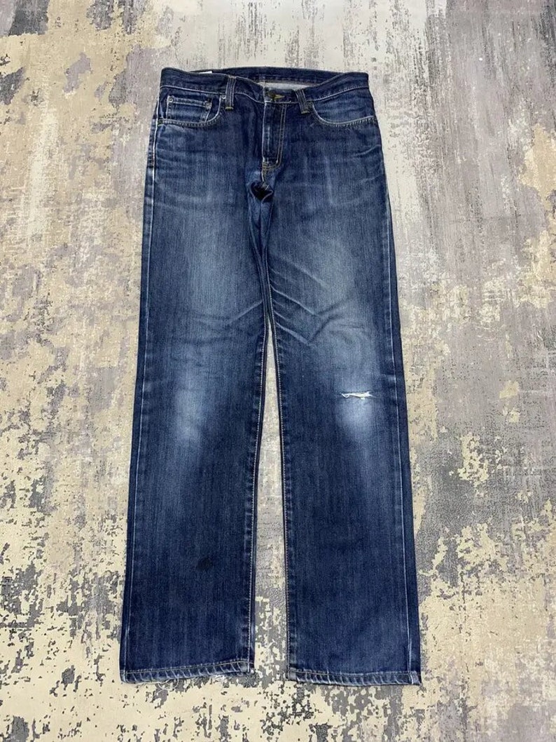 May include: Dark wash denim jeans with a classic five-pocket design. The jeans have a straight leg cut and show signs of wear, including a small tear on the right knee. The denim has faded areas, giving the jeans a vintage look.