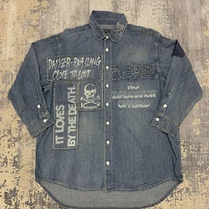 May include: A blue denim button-down shirt featuring various patches and printed text including DANGER-RNA SLANG CLOSE TO LOVE, IT LOVES BY THE DEATH, a skull and crossbones graphic, and the words REBEL NO EXPLANATION OFFERED on the front panels.