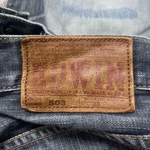 Pode incluir: Close-up de um par de jeans de ganga azul escuro com um remendo de couro castanho. O remendo apresenta a marca "EDWIN" em letras vermelhas, o n&uacute;mero do estilo "503" e o tamanho "36".