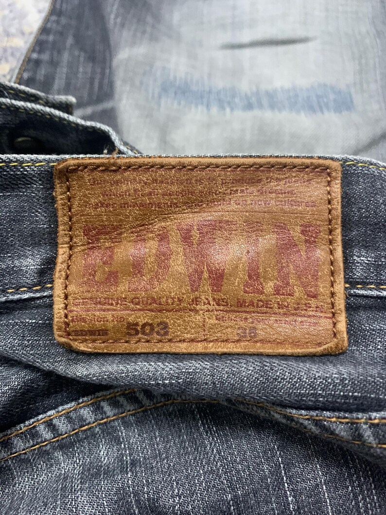 Pode incluir: Close-up de um par de jeans de ganga azul escuro com um remendo de couro castanho. O remendo apresenta a marca "EDWIN" em letras vermelhas, o n&uacute;mero do estilo "503" e o tamanho "36".