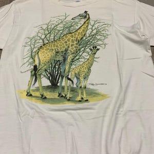 Puede incluir: Camiseta blanca con una ilustraci&oacute;n detallada de dos jirafas, una adulta y una joven, cerca de follaje verde. Las jirafas tienen abrigos estampados marrones y beige. La camiseta est&aacute; hecha de un material suave.