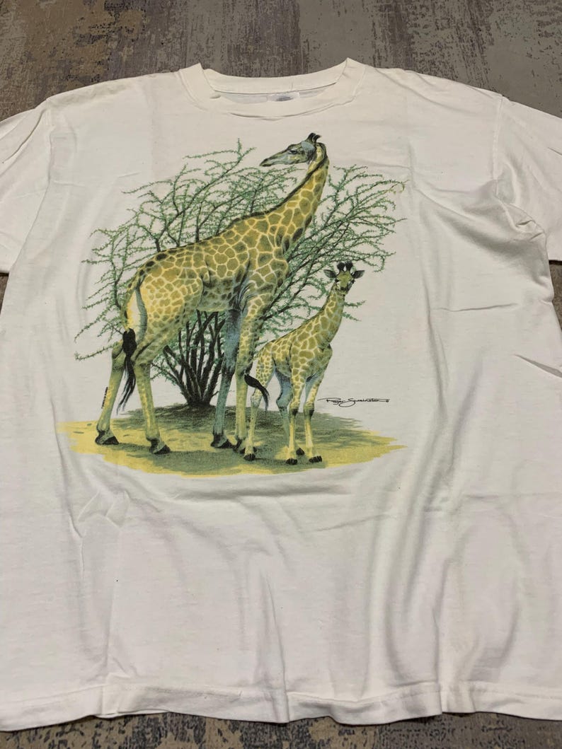 Puede incluir: Camiseta blanca con una ilustraci&oacute;n detallada de dos jirafas, una adulta y una joven, cerca de follaje verde. Las jirafas tienen abrigos estampados marrones y beige. La camiseta est&aacute; hecha de un material suave.
