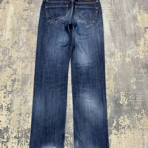 May include: Dark blue denim jeans with a faded wash and distressed details at the hem. The back pockets have a unique design, and a brown leather patch with the brand name "EDWIN" is visible at the waistband.