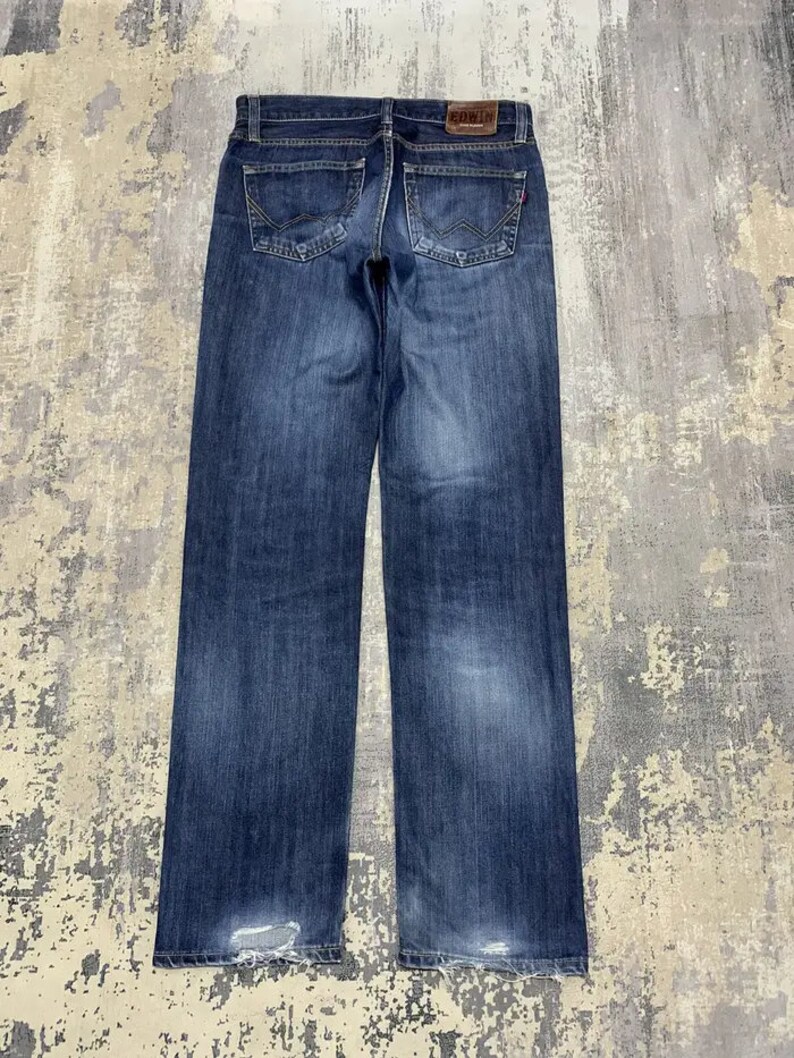 May include: Dark blue denim jeans with a faded wash and distressed details at the hem. The back pockets have a unique design, and a brown leather patch with the brand name "EDWIN" is visible at the waistband.