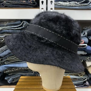 May include: A black, fuzzy bucket hat with a wide black band featuring white stitching. The hat is displayed on a mannequin head. The background shows shelves with folded denim jeans.