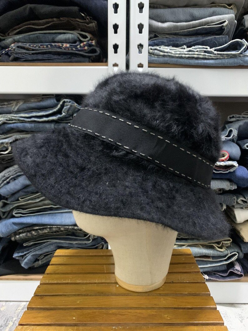 May include: A black, fuzzy bucket hat with a wide black band featuring white stitching. The hat is displayed on a mannequin head. The background shows shelves with folded denim jeans.