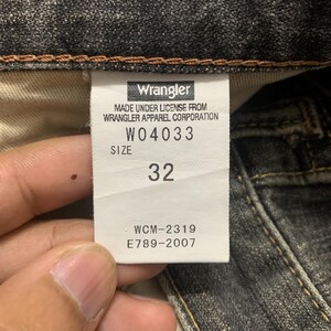 Pu&ograve; includere: Primo piano dell'etichetta di un paio di jeans Wrangler. L'etichetta mostra il nome del marchio, le informazioni sul prodotto e la taglia 32. I jeans sono in denim scuro.