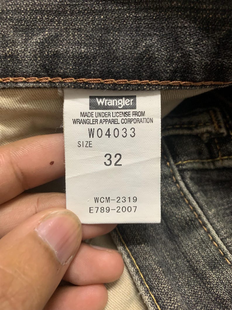 Pu&ograve; includere: Primo piano dell'etichetta di un paio di jeans Wrangler. L'etichetta mostra il nome del marchio, le informazioni sul prodotto e la taglia 32. I jeans sono in denim scuro.