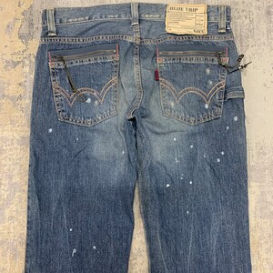 May include: Blue denim jeans with a faded wash and white paint splatters. Features include zippered pockets with pink trim, a red logo tag, and a "BLUE TRIP" label. The jeans have a classic five-pocket design.