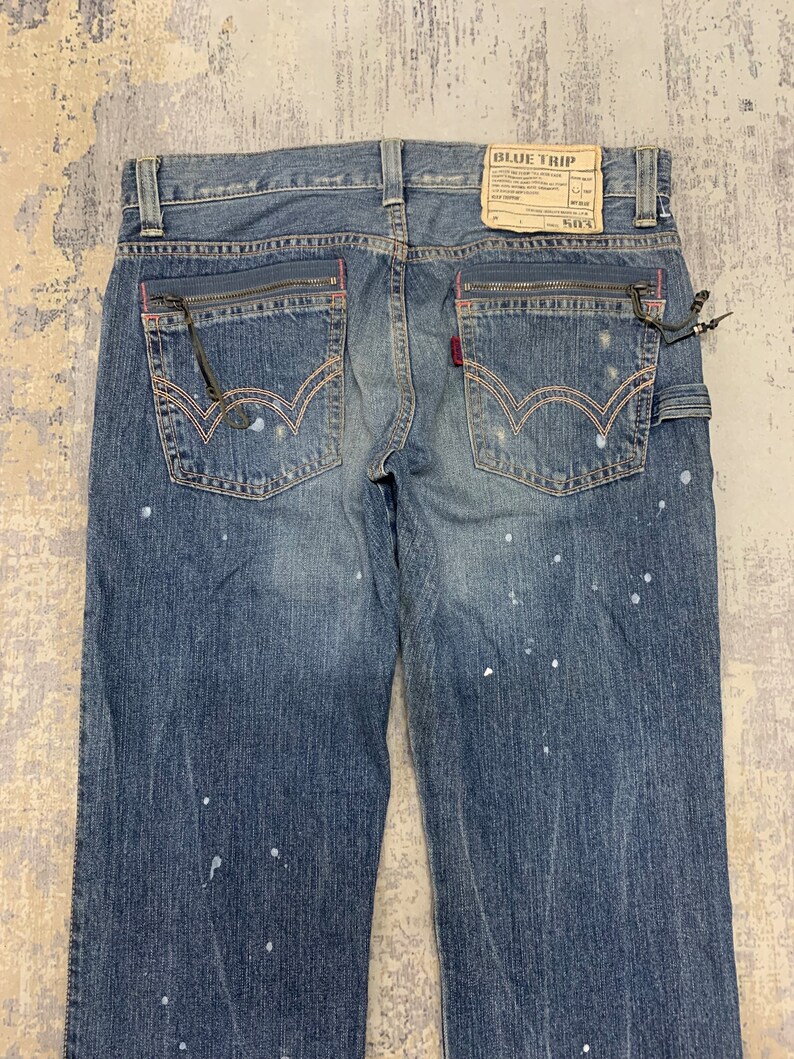 May include: Blue denim jeans with a faded wash and white paint splatters. Features include zippered pockets with pink trim, a red logo tag, and a "BLUE TRIP" label. The jeans have a classic five-pocket design.