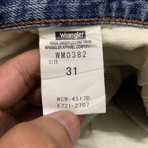 May include: Close-up of a Wrangler jeans tag. The tag displays the brand name, license information, and size 31. The tag also includes the style number WM0382 and other codes: WCM-4517D and E721-2707. The jeans are blue denim.