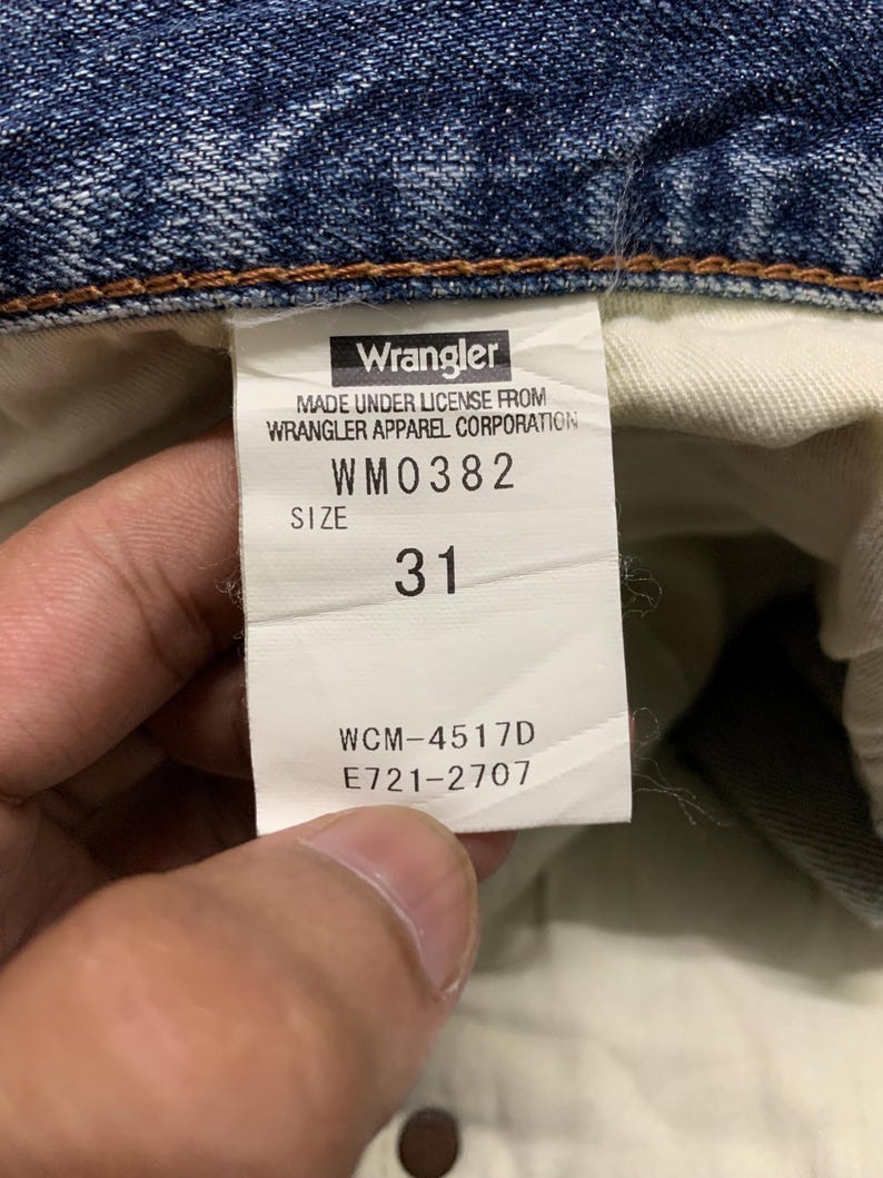 May include: Close-up of a Wrangler jeans tag. The tag displays the brand name, license information, and size 31. The tag also includes the style number WM0382 and other codes: WCM-4517D and E721-2707. The jeans are blue denim.