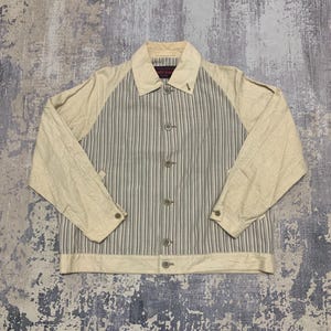 Könnte beinhalten: Beige und grau gestreifte Jacke mit Knopfleiste. Die Jacke hat beige Ärmel und einen Kragen. Die Mittelbahn weist vertikale grau-weiße Streifen auf. Die Jacke hat einen klassischen, lässigen Stil.