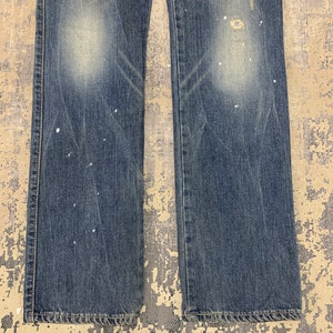 May include: Close-up of blue denim jeans with distressed details. The jeans have a faded wash, visible wear, and white paint splatters. The image shows the lower portion of the jeans, including the hem and stitching.