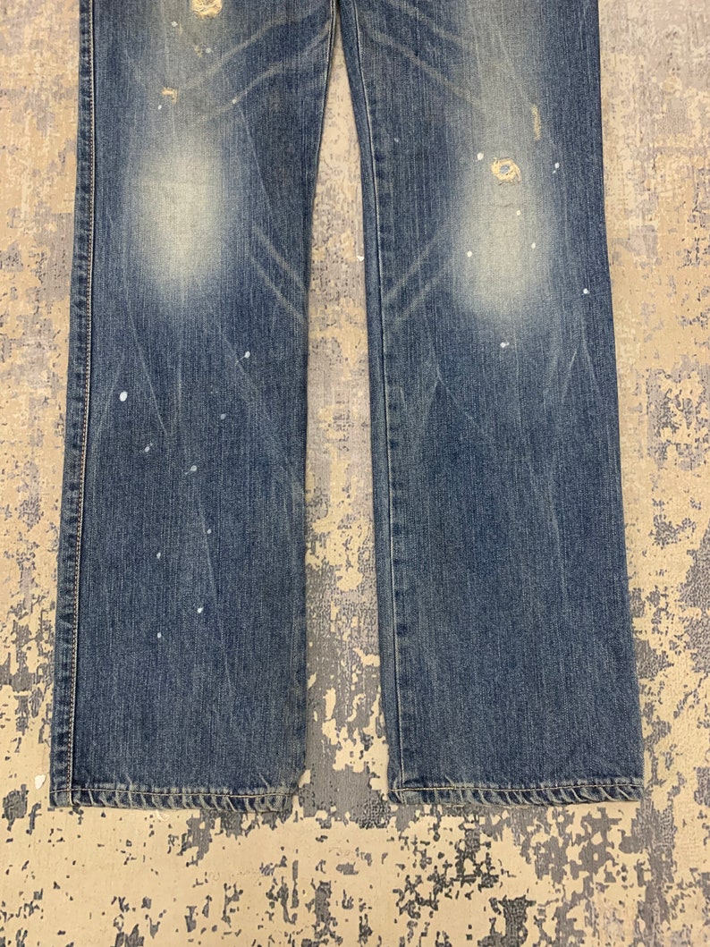 May include: Close-up of blue denim jeans with distressed details. The jeans have a faded wash, visible wear, and white paint splatters. The image shows the lower portion of the jeans, including the hem and stitching.
