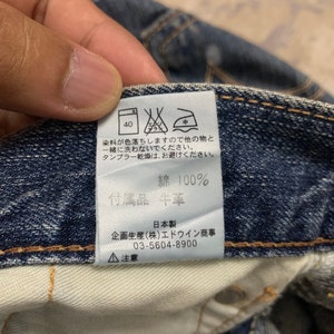 May include: Close-up of a denim garment's interior, showcasing a care label with Japanese text and symbols. The label includes washing instructions, material composition (100% cotton), and manufacturer details. The denim is a deep blue color.