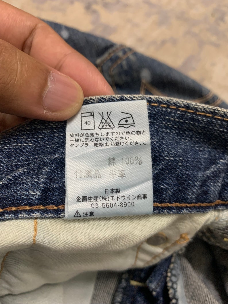 May include: Close-up of a denim garment's interior, showcasing a care label with Japanese text and symbols. The label includes washing instructions, material composition (100% cotton), and manufacturer details. The denim is a deep blue color.