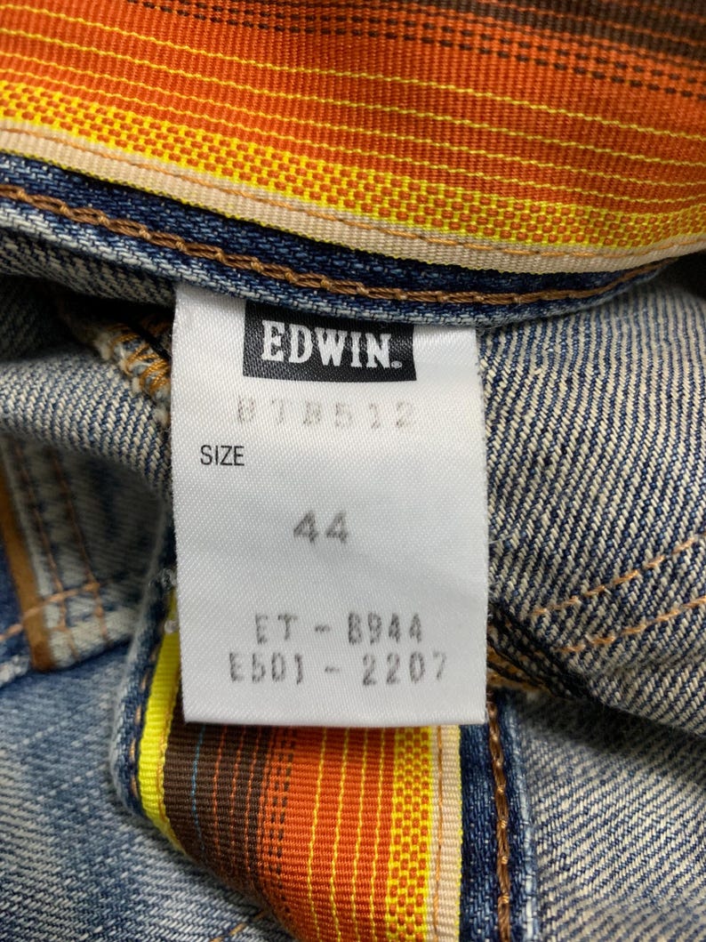 Może przedstawiać: Zbliżenie na element odzieży dżinsowej z metką marki EDWIN. Na metce widnieje rozmiar 44 oraz inne kody produktu. Wewnętrzna podszewka ma kolorowy wz&oacute;r w paski w odcieniach pomarańczu, ż&oacute;łci i brązu.