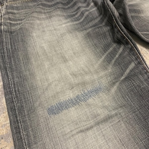 Pode incluir: Close-up de cal&ccedil;as de ganga cinzentas com lavagem desbotada. As cal&ccedil;as de ganga t&ecirc;m um design cl&aacute;ssico de cinco bolsos e fecho de bot&atilde;o e z&iacute;per. Uma mancha de costura azul &eacute; vis&iacute;vel na frente, indicando um reparo ou elemento de design.