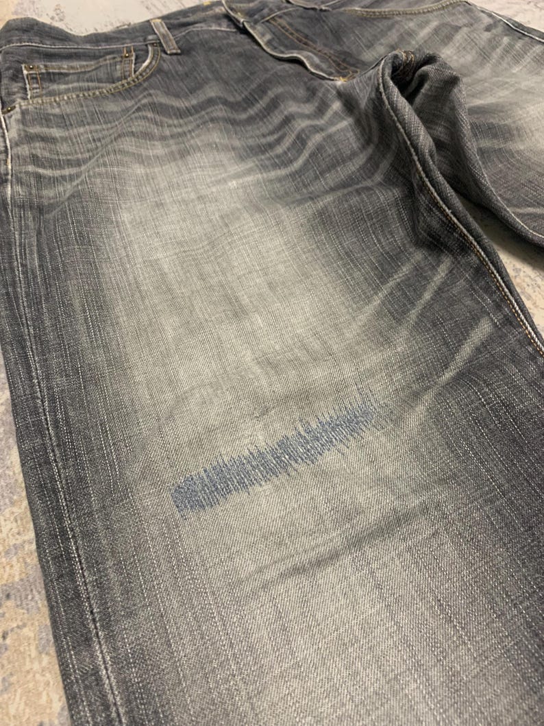 Pode incluir: Close-up de cal&ccedil;as de ganga cinzentas com lavagem desbotada. As cal&ccedil;as de ganga t&ecirc;m um design cl&aacute;ssico de cinco bolsos e fecho de bot&atilde;o e z&iacute;per. Uma mancha de costura azul &eacute; vis&iacute;vel na frente, indicando um reparo ou elemento de design.