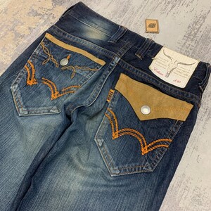 May include: Dark wash denim jeans with tan suede pocket accents and orange stitching. The back pockets feature a unique embroidered design. The Edwin brand label is visible on the back pocket. A small leather patch is also present.