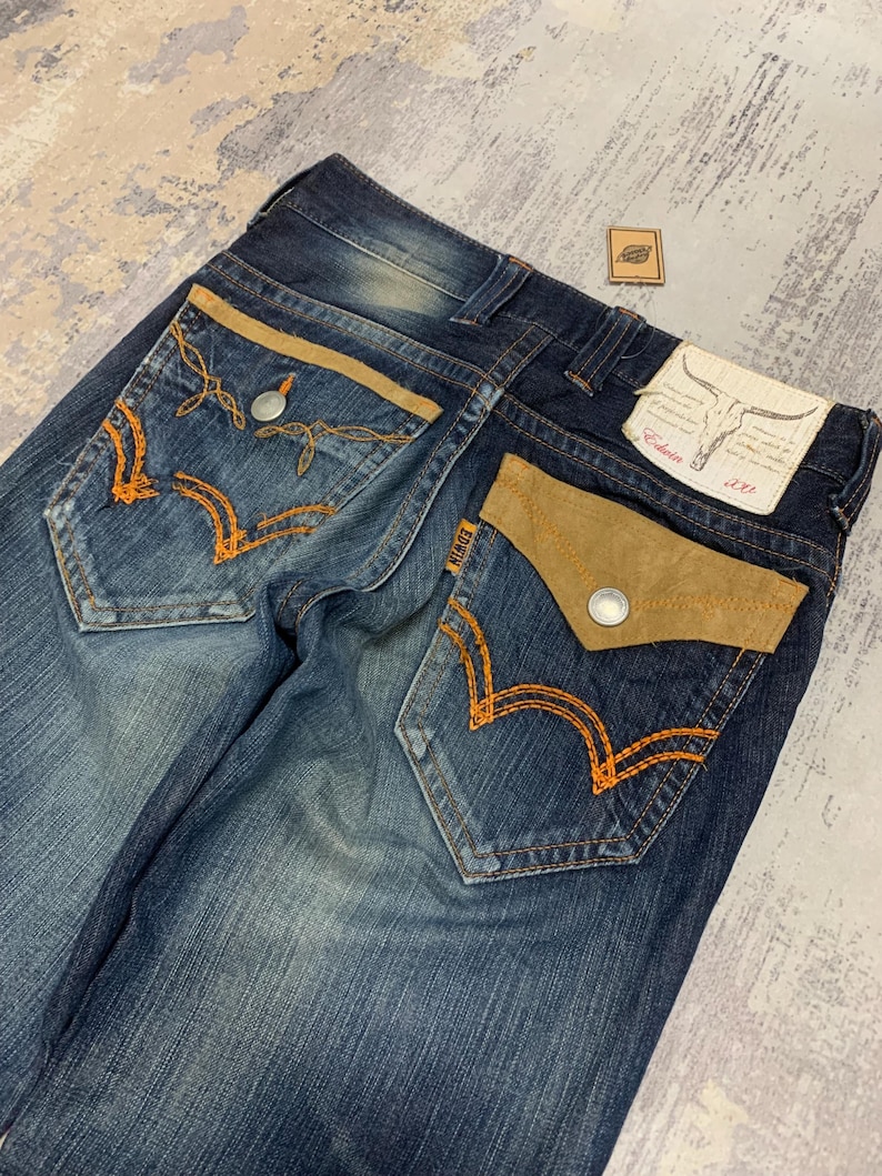 May include: Dark wash denim jeans with tan suede pocket accents and orange stitching. The back pockets feature a unique embroidered design. The Edwin brand label is visible on the back pocket. A small leather patch is also present.
