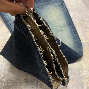 May include: Close-up of blue denim jeans with a distressed, worn look. The image highlights the frayed edges and multiple layers of the denim fabric, showcasing the inside and outside textures. The jeans have a faded wash and a classic five-pocket design.