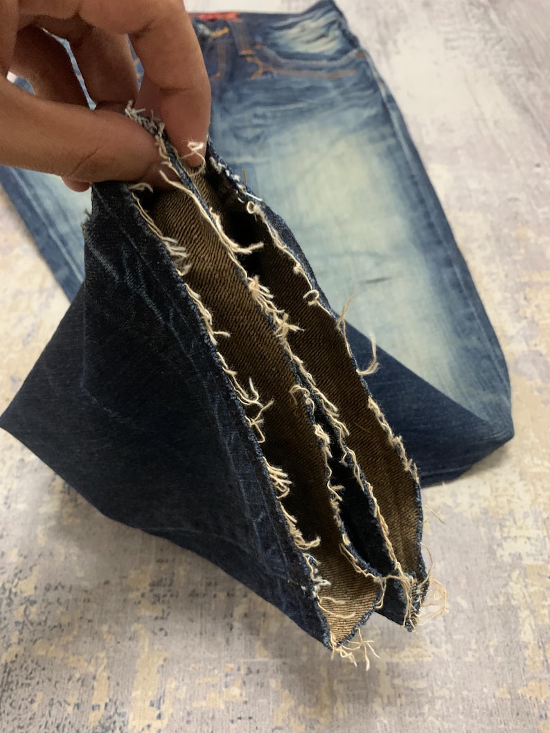May include: Close-up of blue denim jeans with a distressed, worn look. The image highlights the frayed edges and multiple layers of the denim fabric, showcasing the inside and outside textures. The jeans have a faded wash and a classic five-pocket design.