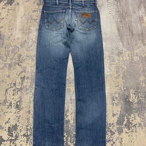 May include: Blue denim jeans with the Wrangler logo on the back pockets. The jeans have a classic five-pocket design and a brown leather patch. The jeans are a medium wash and appear to be pre-worn.