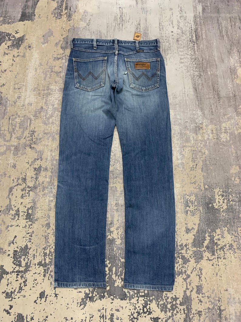 May include: Blue denim jeans with the Wrangler logo on the back pockets. The jeans have a classic five-pocket design and a brown leather patch. The jeans are a medium wash and appear to be pre-worn.