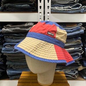 Puede incluir: Un sombrero de pescador con bloques de color que presenta paneles de tela roja, beige y azul. El sombrero tiene una banda azul alrededor del centro y un parche rectangular negro en la parte frontal con la palabra BREEZE en letras blancas.