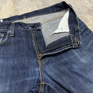 May include: Close-up of dark blue denim jeans with a partially unzipped brass zipper. The jeans have a button closure and visible stitching in a contrasting yellow-orange thread. The interior lining is a light color.