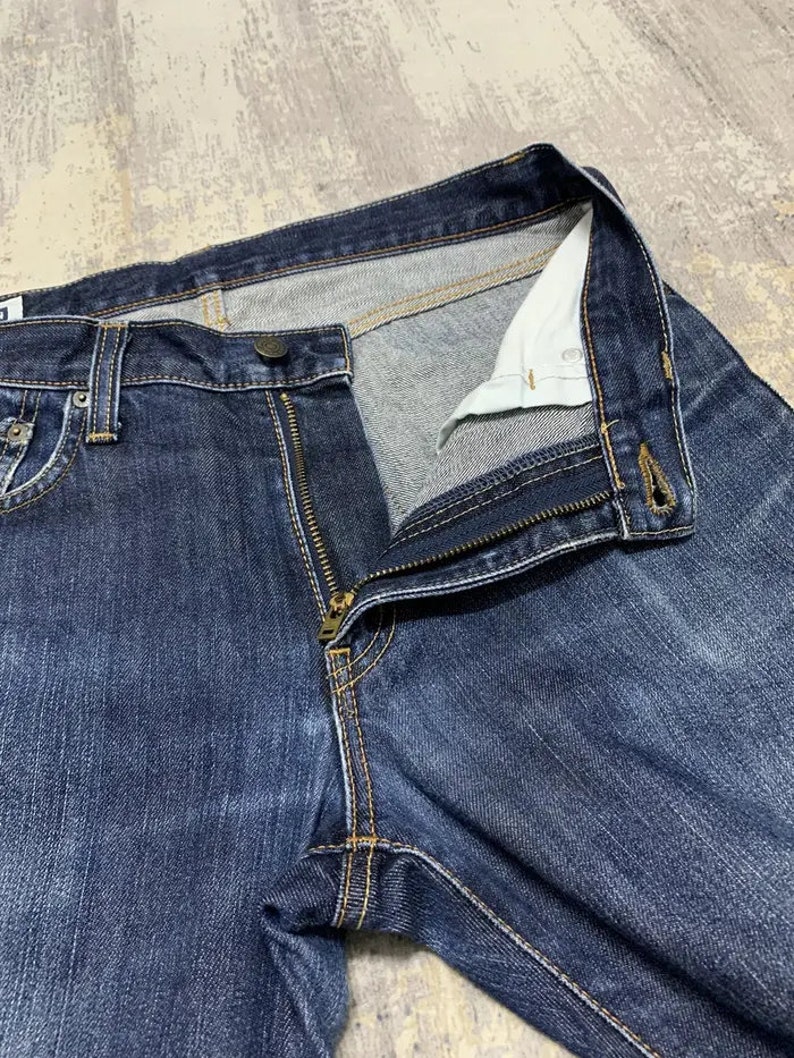 May include: Close-up of dark blue denim jeans with a partially unzipped brass zipper. The jeans have a button closure and visible stitching in a contrasting yellow-orange thread. The interior lining is a light color.