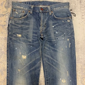 May include: Faded blue denim jeans with distressed details, including worn patches and paint splatters. The jeans have a classic five-pocket design with a button and zipper closure. The denim has a vintage wash.