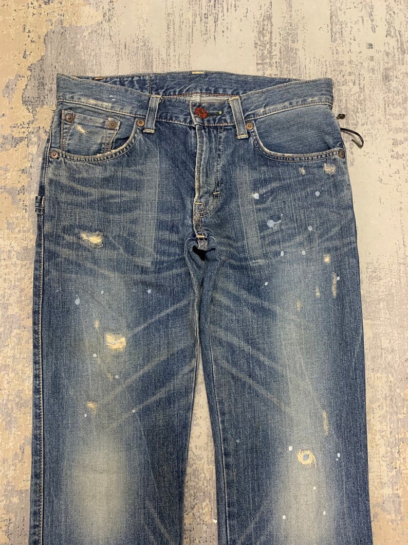 May include: Faded blue denim jeans with distressed details, including worn patches and paint splatters. The jeans have a classic five-pocket design with a button and zipper closure. The denim has a vintage wash.