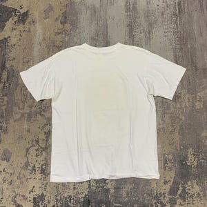 Puede incluir: Una camiseta blanca lisa, colocada sobre una superficie gris texturizada. La camiseta tiene mangas cortas y cuello redondo. La tela parece suave y la camiseta est&aacute; limpia y sin usar.