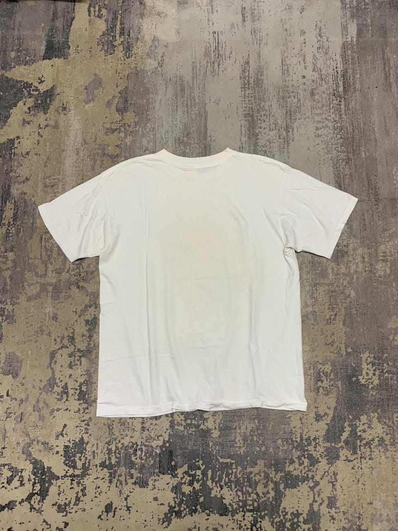 Puede incluir: Una camiseta blanca lisa, colocada sobre una superficie gris texturizada. La camiseta tiene mangas cortas y cuello redondo. La tela parece suave y la camiseta est&aacute; limpia y sin usar.
