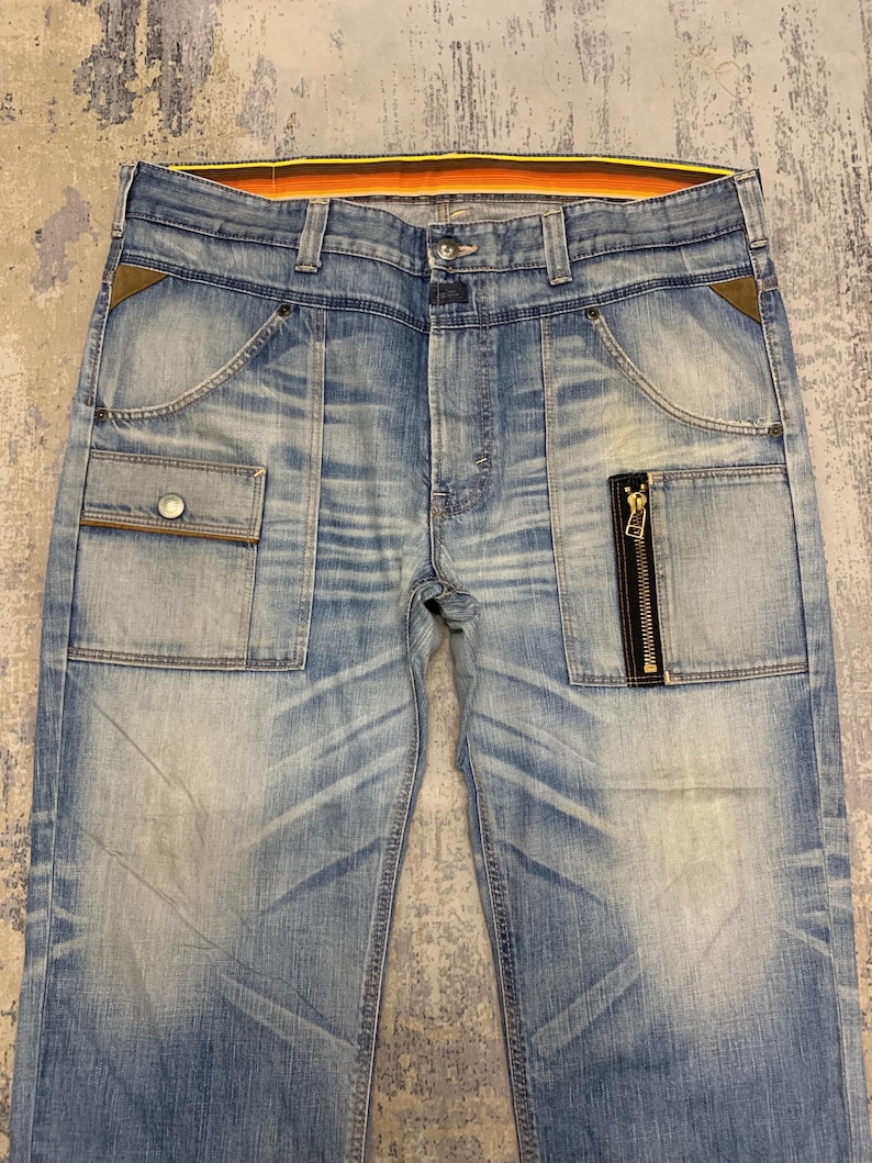 May include: Light blue denim jeans with multiple pockets, including a cargo pocket and a zippered pocket. The jeans have a faded wash and a contrasting orange and yellow waistband. The jeans have a button and zipper closure.