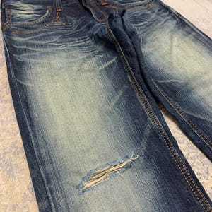 May include: Close-up of distressed blue denim jeans. The jeans have a faded wash, a ripped knee, and visible stitching. The denim is a medium weight, and the jeans appear to be pre-worn.