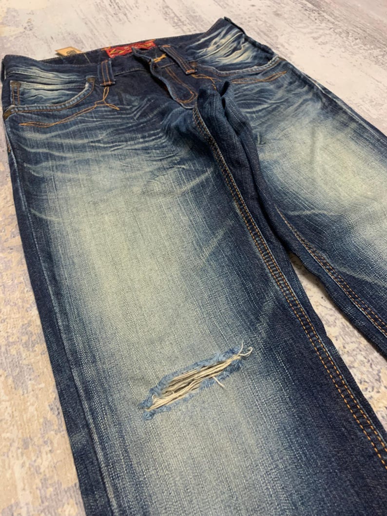 May include: Close-up of distressed blue denim jeans. The jeans have a faded wash, a ripped knee, and visible stitching. The denim is a medium weight, and the jeans appear to be pre-worn.