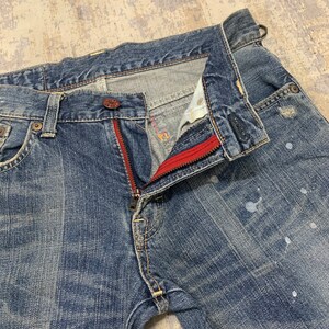 May include: Close-up of blue denim jeans with a partially unzipped fly. The zipper has a red accent, and there are white paint splatters on the denim. The jeans have a classic five-pocket design.