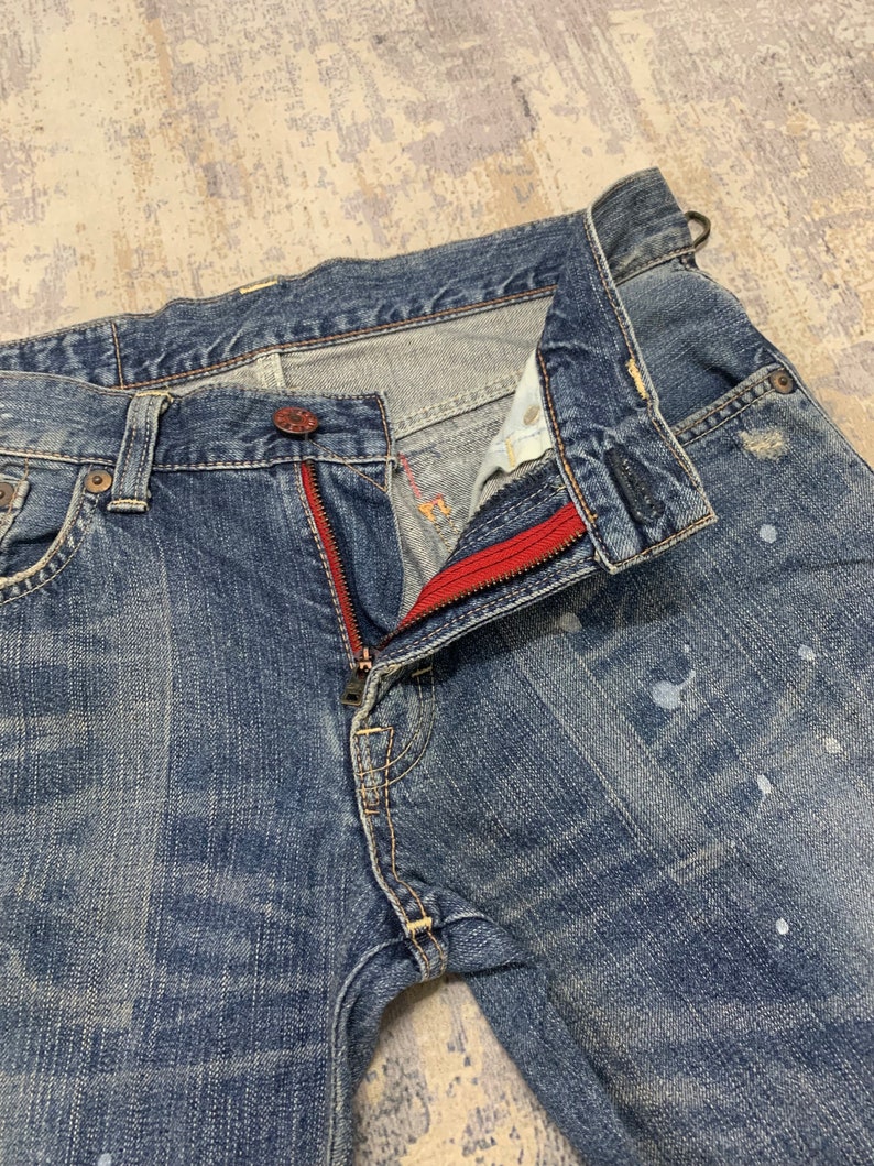 May include: Close-up of blue denim jeans with a partially unzipped fly. The zipper has a red accent, and there are white paint splatters on the denim. The jeans have a classic five-pocket design.