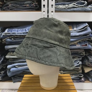 Pode incluir: Um chapéu bucket verde-oliva escuro com um acabamento de tecido texturizado e ligeiramente desgastado. O chapéu apresenta uma copa arredondada e uma aba inclinada para baixo, construído com costuras visíveis ao longo das bordas.